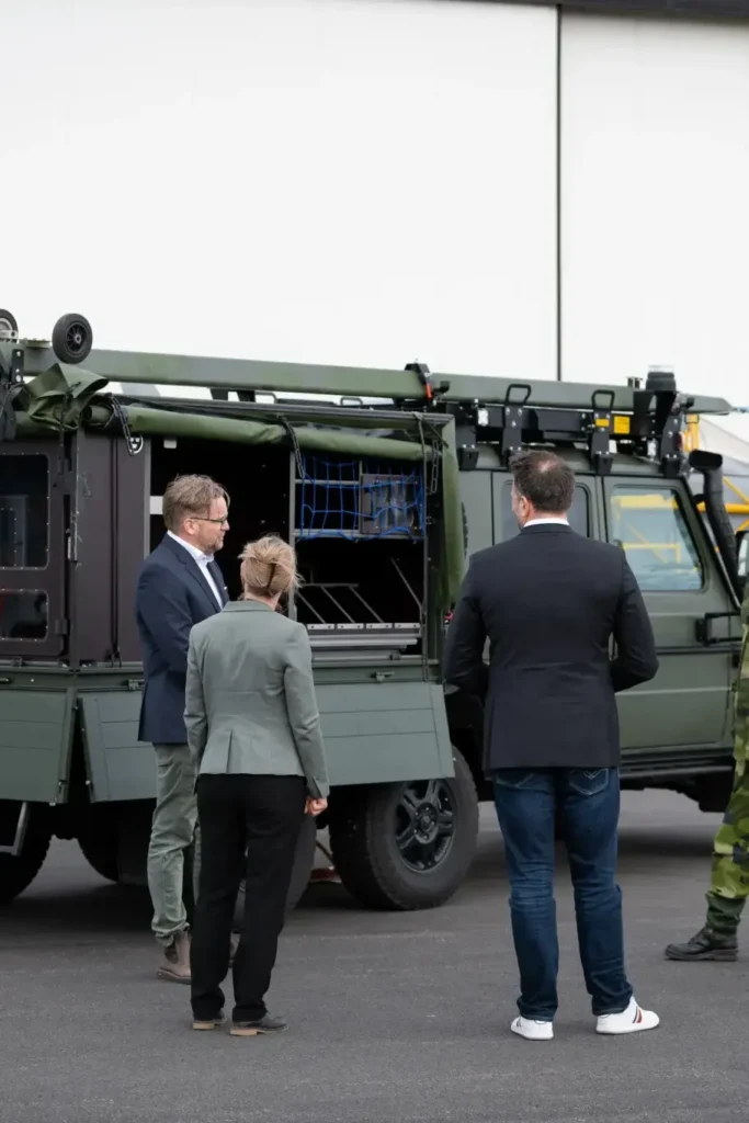 Bild på två män och en kvinna framför ett militärfordon.